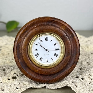 May include: A small, round quartz clock with a dark brown wooden frame and a gold-colored bezel. The clock face is white with black Roman numerals and hands. The clock is displayed on a cream-colored, crocheted doily.