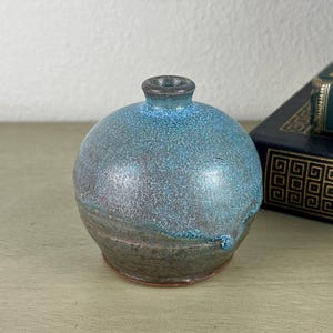 May include: A small, spherical ceramic vase with a narrow neck and a textured glaze. The upper portion features a vibrant, speckled blue finish that transitions into a darker, earthy brown tone toward the base, creating a distinct color gradient.