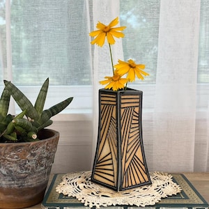 May include: A square-shaped vase with a light tan surface and black geometric line patterns, holding two yellow flowers. Next to it is a green potted plant in a textured brown pot, both resting on a dark green book with gold accents.