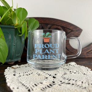 May include: Clear glass mug with the words "PROUD PLANT PARENT" in light blue. A small illustration of a potted plant is above the text. The mug has a handle and is sitting on a white lace doily.