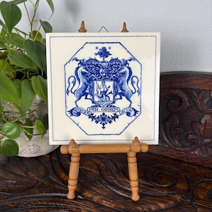 May include: A square, white ceramic tile with a blue heraldic design, displayed on a small wooden easel. The design features a coat of arms with lions, a shield, and the text "PER ORBEM DICOR". The easel is made of light brown wood.