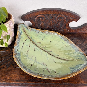 May include: A ceramic leaf-shaped dish with a light green glaze and a dark brown vein pattern. The dish has a golden-brown rim and sits on a dark wooden surface. A potted plant is visible in the background.