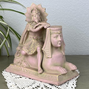 May include: A pink and gold Egyptian-themed figurine featuring a pharaoh figure leaning on a sphinx. The figurine has intricate details and a vintage aesthetic. The pharaoh wears a headdress and cape, while the sphinx has a serene expression.