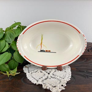 May include: Cream-colored oval serving dish with a red rim and a decorative design. The dish features a minimalist illustration of a plant and train. The dish is displayed on a white lace doily.