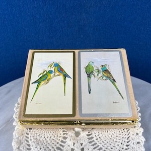 May include: A vintage playing card box featuring two sets of colorful bird illustrations. The box has a gold trim and a beige exterior. Each card set depicts two birds perched on a branch, with a blue background.