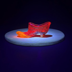 May include: A red glass shoe with a patterned design. The shoe is on a white circular platform.