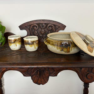 May include: A wooden table with a carved design features a brown and white ceramic serving dish with a lid and two smaller ceramic cups.