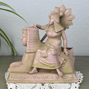 May include: A pink and green Egyptian-themed figurine featuring a seated figure atop a sphinx. The figure wears a headdress and traditional garb. The base is inscribed with hieroglyphs. The figurine is displayed on a white lace doily.