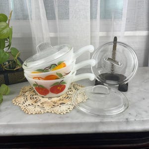 May include: A stack of three white milk glass bowls with handles, each decorated with orange and green fruit illustrations. The bowls are topped with a clear glass lid, and two additional clear glass lids are placed nearby on the surface.