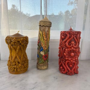 May include: Three decorative pillar candles. The candle on the left is yellow with floral and braided designs. The middle candle is tan with wheat and floral accents. The candle on the right is red with an ornate, swirling pattern.