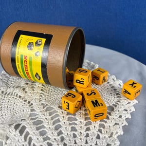 May include: A vintage Scribbage word game set. The brown cardboard container is tipped over, spilling out yellow letter dice with black lettering. The container has a yellow and black label with the game's name.