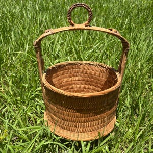 May include: A round, light brown woven basket featuring a tall, arched handle with a small, circular loop at the very top. The basket has a cylindrical body with a flat base and a reinforced rim, constructed from thin, flexible strips of natural material.