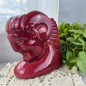 May include: A burgundy ceramic head sculpture with ram horns. The sculpture is placed on a cream-colored, crocheted doily. Green plants are visible in the background. The sculpture has a glossy finish and detailed facial features.