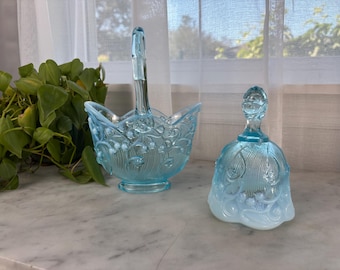 May include: A light blue glass basket with a handle and a matching bell. The basket has a floral design and the bell has a detailed top. Both items are on a marble surface.