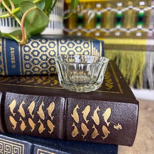May include: A clear glass dish with a fluted rim sits on top of two leather-bound books. The books have gold embossed designs on the covers.