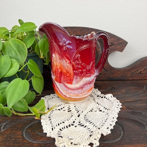 May include: A vintage red glass jug with a handle, featuring a floral design and a ruffled rim. The jug sits on a cream-coloured, heart-shaped doily. A green plant is visible in the background.