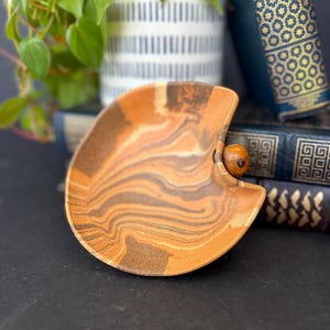 May include: A small, brown and white ceramic dish with a swirl pattern. The dish has a wooden knob on the side and is sitting on top of a stack of books.