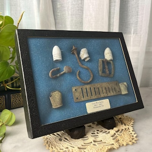 May include: A black rectangular display frame containing various metal artifacts, including bullets, a hook, a buckle, and a slotted plate. A small label reads Civil War Relics 1861-1865 Richmond, VA. The items are mounted against a blue background.