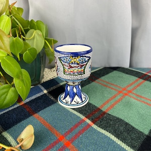 May include: A ceramic goblet with a mosaic design. The goblet features a blue rim and a detailed pattern of blue, red, yellow, and white tiles. The base is white with blue accents, and the goblet sits on a plaid blanket.