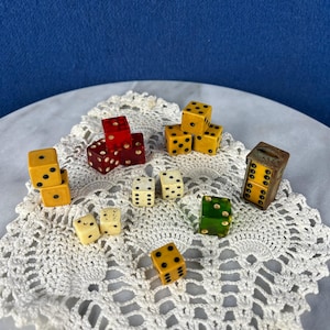 May include: A collection of vintage dice in various colors, including yellow, red, green, and cream. The dice are arranged on a white, lace doily against a blue background. Some dice are stacked, and others are scattered.