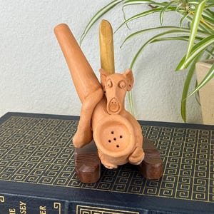 May include: A terracotta-colored sculpture of a dog-like creature with a large bowl-shaped body and a long, angled spout. The sculpture rests on a small, dark brown wooden base, with a wooden stick extending upwards. The piece is set against a neutral background.
