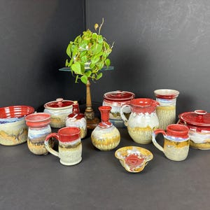 May include: A collection of handmade pottery in various shapes and sizes, including mugs, bowls, and pitchers. The pottery is decorated with red, white, and brown glazes, and features a rustic, earthy aesthetic.