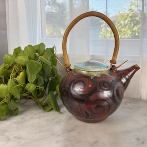 May include: A handcrafted ceramic teapot with a red and black swirl design. It features a light blue lid and a woven handle. The teapot is approximately 8 inches tall.