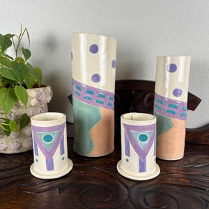May include: A collection of four ceramic vases and cups with geometric designs. The vases are tall and cylindrical, featuring a cream base with purple polka dots, teal, and peach accents. The cups have a similar design.