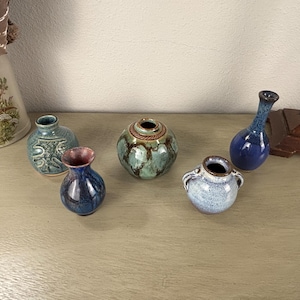 May include: Five small ceramic vases in different shapes and colours. The vases feature shades of blue, green, and brown, with textured and glazed surfaces. One vase has a flared rim with a reddish-brown interior.