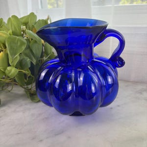 May include: A cobalt blue glass pitcher with a rounded, pumpkin-like body and a curved handle. The pitcher has a spout for easy pouring and is displayed on a marble surface. The glass reflects light, highlighting its shape and color.