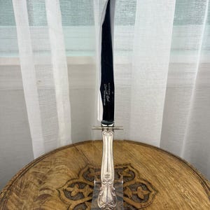 May include: A silver-toned knife with a curved, polished stainless steel blade and an ornate, embossed metal handle. The blade features the engraved text "Made in Sheffield England" and "Stainless Steel". The knife is displayed vertically.