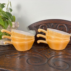 May include: Two stacks of vintage orange glass soup bowls with handles. The bowls have a ribbed texture and a light orange color. The bowls are on a dark wooden surface, with a plant and decorative wooden piece in the background.