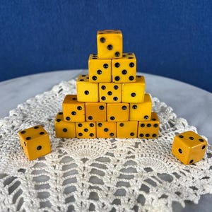 May include: A pyramid of yellow dice with black dots, arranged on a white lace doily. Two additional dice are placed near the pyramid. The background is a gradient of blue.