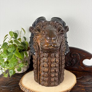 May include: A brown and black sculpted ram head figurine featuring detailed curled wool textures and prominent curved horns. The sculpture has a dark brown face with defined facial features and a vertical pattern of circular swirls along the neck.