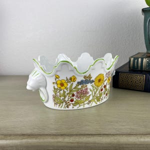 May include: A white ceramic oval bowl with a scalloped rim and a green painted edge. The exterior features a colorful botanical pattern of yellow, pink, and blue wildflowers. The bowl has a sculpted animal head handle on the side.