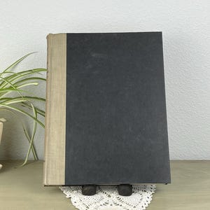 May include: A hardcover book with a black cover and a tan spine. The book is displayed on a white lace doily and held up by two small, dark wooden bookends. The book is standing upright on a light green surface.