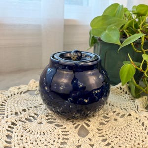 May include: A small, round, dark blue ceramic jar with a lid, speckled with lighter blue and white. The jar sits on a cream-colored, crocheted doily. A green potted plant is in the background.