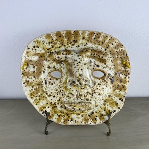 May include: A ceramic wall mask featuring a stylized human face with a cream-colored base and brown speckled glaze. The mask has cut-out eye openings, a prominent nose, and a small mouth, with yellow cords attached to the sides for hanging.