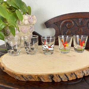 May include: Five clear shot glasses with various rooster and bird designs. The glasses are arranged on a wooden slice, with a plant and carved wooden background. The rooster designs are in red, black, and white.