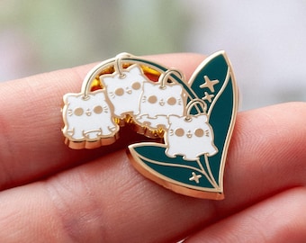 May include: A gold-toned enamel pin featuring three white cat faces hanging from a gold arch, resembling lily of the valley flowers. The pin has a dark green leaf with gold accents and small star details. The pin is approximately 1 inch in length.
