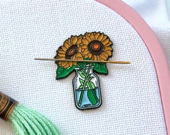 May include: A metal needle minder featuring a sunflower bouquet in a glass jar, attached to white Aida fabric within a pink Nurge embroidery hoop. Three skeins of embroidery floss in orange, peach, and light green are positioned across the fabric.
