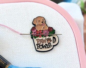 May include: A metal enamel needle minder featuring a golden dog sitting in a mug filled with pink and red flowers. The mug has the text PAW POWER printed on the front. A silver needle is attached to the minder, which is placed on white fabric.
