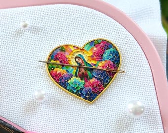 May include: A heart-shaped needle minder featuring a colorful illustration of the Virgin Mary surrounded by vibrant flowers, attached to white Aida fabric in a light pink Nurge embroidery hoop with pink and light green embroidery floss skeins.