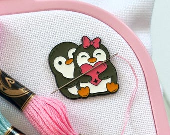May include: A light pink Nurge embroidery hoop holding white fabric, featuring a metal enamel pin of two penguins holding a pink heart. Two skeins of embroidery floss in light pink and light blue are placed across the fabric with a needle.