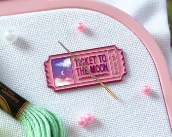 May include: A light pink Nurge embroidery hoop holding white fabric, featuring a rectangular enamel pin shaped like a ticket with the text Ticket to the Moon, a gold needle, two skeins of light green embroidery thread, and scattered small beads.
