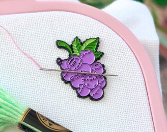 May include: A pink Nurge embroidery hoop holding white fabric with a purple grape-shaped enamel pin featuring cat faces. A silver needle with pink thread pierces the fabric. Two skeins of embroidery floss in light green and blue are visible.