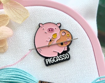 May include: A pink enamel pin shaped like a pig holding a paintbrush and a paint palette, with the text PIGCASSO printed in black at the bottom. The pin is attached to white fabric stretched in a pink embroidery hoop with light blue and lime green thread.