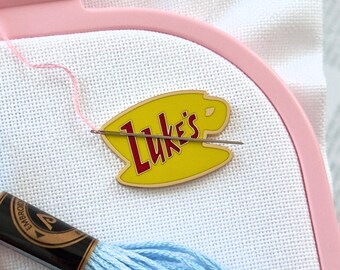 May include: A light pink plastic embroidery hoop holding white fabric, featuring a yellow enamel pin shaped like a coffee cup with the red text Luke's. A silver needle is threaded with pink floss, alongside two skeins of blue and pink embroidery thread.