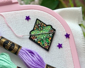 May include: A light pink Nurge embroidery hoop holding white fabric, featuring a diamond-shaped enamel pin with a green moth and floral design, a silver needle with pink thread, two skeins of embroidery floss in purple and mint green.