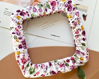 Puede incluir: Una diadema blanca con estampado floral de forma cuadrada. La diadema presenta un patrón de flores moradas, rosas y amarillas con hojas verdes. La palabra «SIMPLE» es visible en una revista en el fondo.